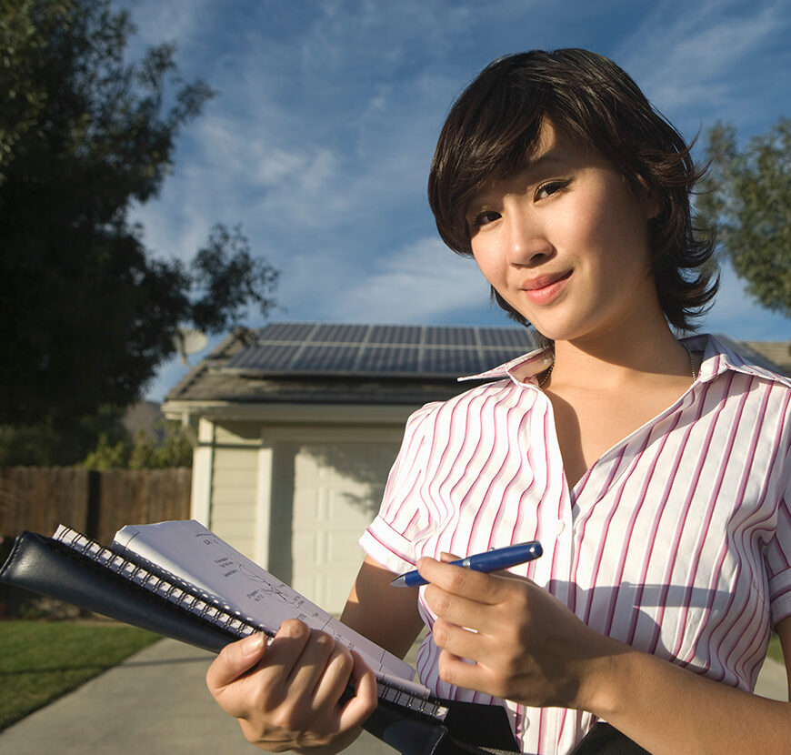 A female appraiser stands outside a home with solar panels.